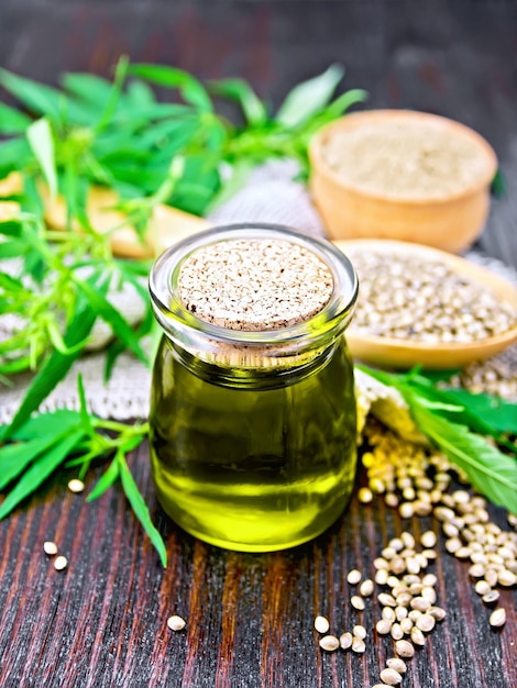 Hemp oil in a glass jar grain in a spoon and flour in a bowl on a napkin of burlap leaves and stalks of cannabis against a dark wooden board