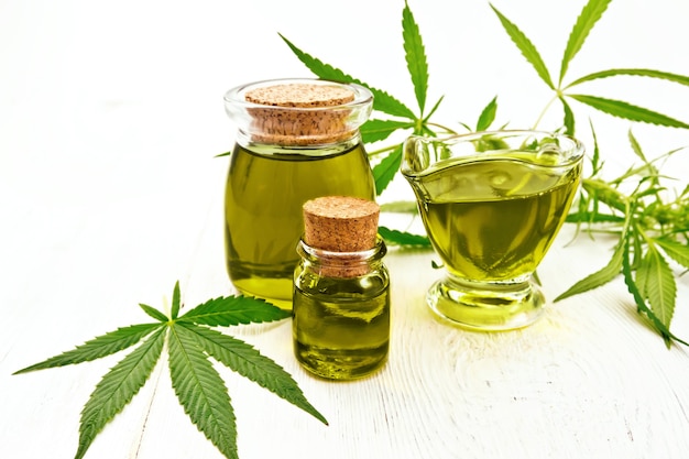 Hemp oil in two glass jars and sauceboat, leaves and stalks of cannabis on the background of light wooden boards