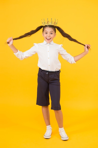 I suoi capelli sono il suo lusso. piccola ragazza felice in corona che tiene i capelli lunghi del brunette su sfondo giallo. acconciare i capelli per il ballo di fine anno nel salone di bellezza. fare un'acconciatura fantasia con i capelli lunghi.