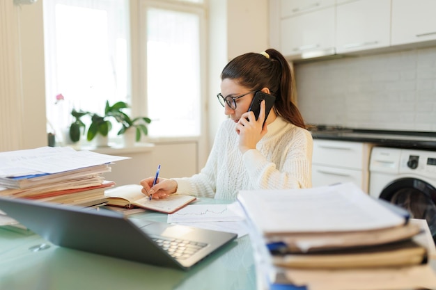 Photo in her home workspace a woman takes a break from laptop work talking on the phone while surrounded