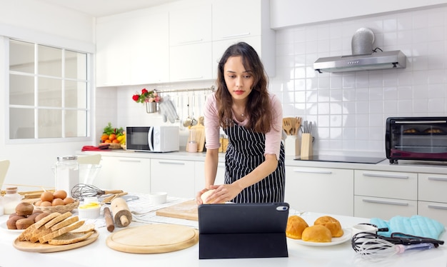 In her kitchen at home, a lovely young woman is taking an online cooking class on a tablet computer.