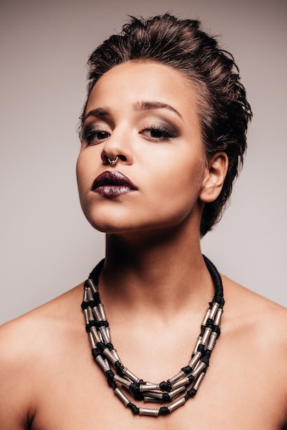 In her own style. Portrait of beautiful young shirtless woman with nose ring and necklace looking at camera while standing against grey background