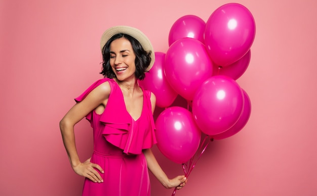 Photo her perfect birthday. young charming woman in a fuchsia dress and straw hat is holding pink balloons in her left hand and laughing.