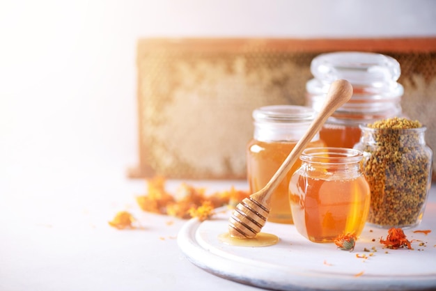 Травяной мед в банке с ковшом, пчелиные соты, пыльца, гранулы, цветы календулы на сером фоне