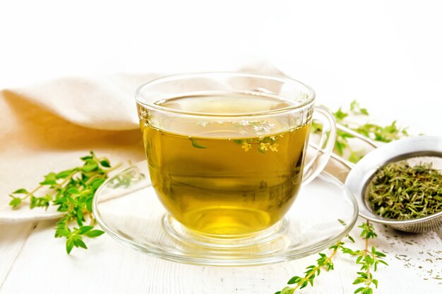 Herbal tea in a glass cup of thyme, a metal strainer with dry leaves, a linen napkin against a white wooden board