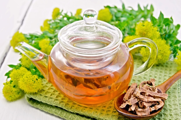 Photo herbal tea in a glass teapot on a napkin, spoon with dry roots of rhodiola rosea, fresh flowers rhodiola rosea on the background light wooden boards
