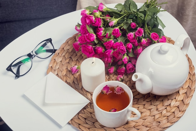 Tisana, rose rosa, candela accesa sul tavolino bianco.