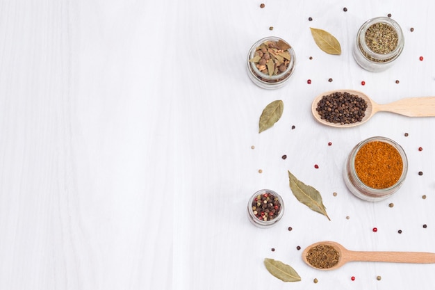 Herbs and spices in wooden spoons and glass jars on white wooden table. Top view