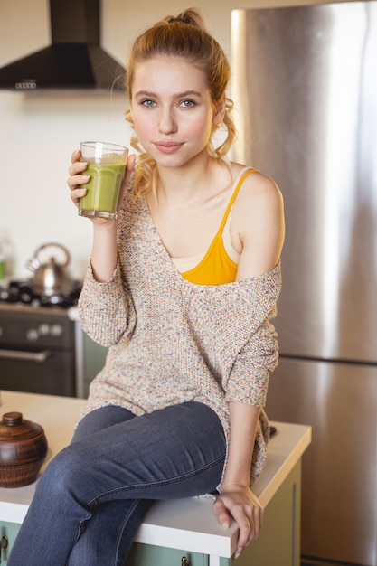 Here I am. Delighted girl sitting on the table in kitchen while drinking smoothie