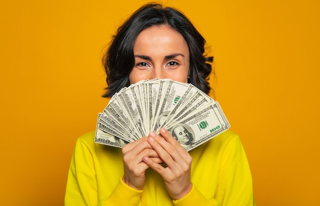 Here is my salary! a young girl in a yellow hoodie, smiling with  her eyes, hiding her face behind big amount of money in her hands.