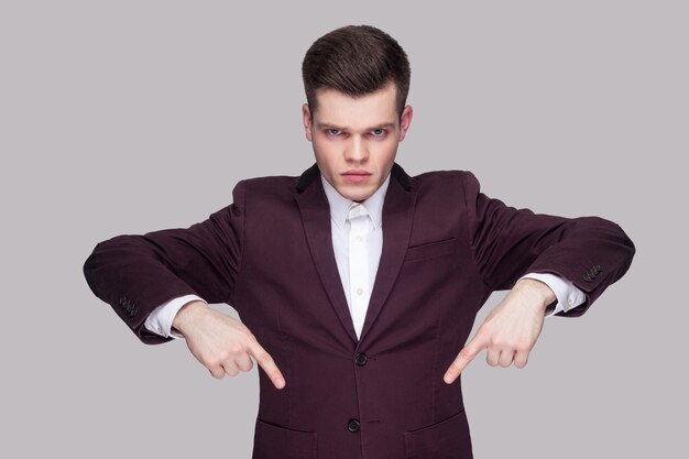 Here and right now. Portrait of serious handsome young man in violet suit, white shirt, standing, looking at camera and pointing down with raised arms. indoor studio shot, isolated on grey background.