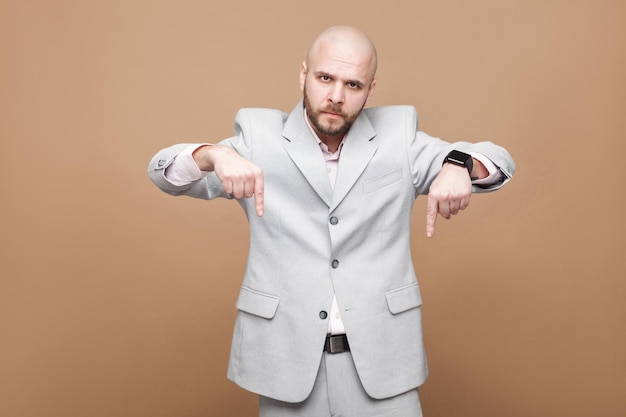 Here and right now. Serious middle aged bald bearded businessman boss in classic light gray suit standing, looking at camera and showing pleace. indoor studio shot, isolated on light brown background