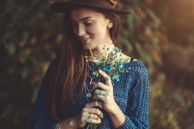 Herfst brunette boho chique vrouw in een bruine hoed en gebreide blauwe trui met boeket van wilde bloemen in handen in de herfst bos buiten in de herfst
