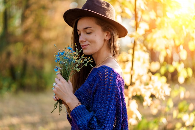 Herfst brunette vrouw met lang haar in een bruine hoed en in een gebreide blauwe trui geniet van de geur van wilde bloemen in de herfst bos buiten in de herfst