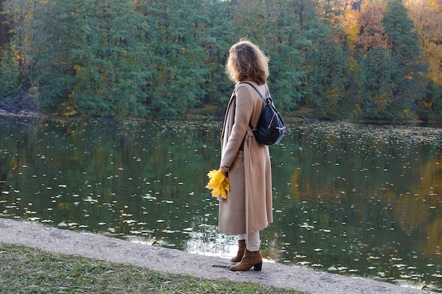 Herfst. Een jonge vrouw staat op de oever van het meer en kijkt in de verte. Ze is gekleed in trendy zakelijke kleding in beige. In haar handen heeft ze een boeket esdoornbladeren.