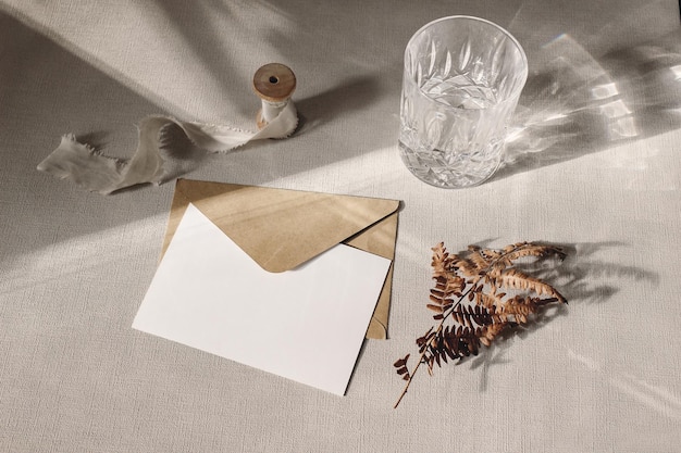 Herfst herfst briefpapier mockup scène lege wenskaart uitnodiging ambachtelijke papieren envelop droog varenblad en sprankelend glas water op grijze tafellinnen achtergrond in zonlicht hard schaduwspel
