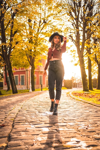 Herfst levensstijl bij zonsondergang blonde blanke vrouw in een rode trui en zwarte hoed genieten van de natuur in een park met bomen wandelen op het prachtige pad van het stadspark