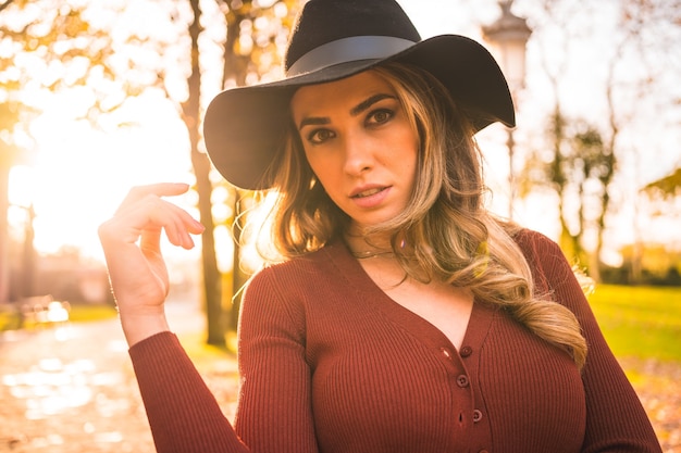 Herfst levensstijl bij zonsondergang blonde blanke vrouw in een rode trui en zwarte hoed genieten van de natuur in een park met bomen wandelen op het prachtige pad van het stadspark