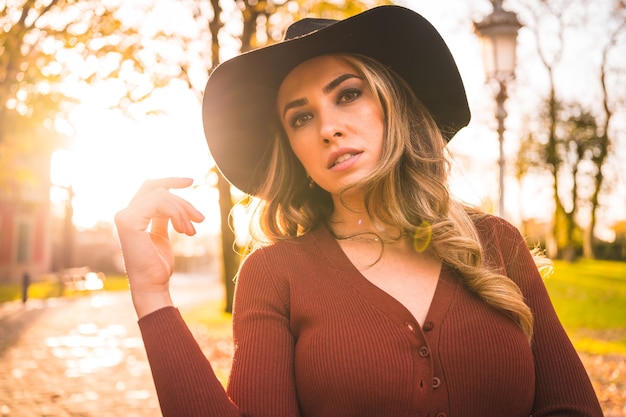 Herfst levensstijl bij zonsondergang blonde blanke vrouw in een rode trui en zwarte hoed genieten van de natuur in een park met bomen wandelen op het prachtige pad van het stadspark