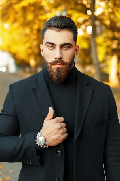 Herfst portret van een hipster man met een baard en snor in een stijlvolle zwarte jas en een horloge op een zonnige dag