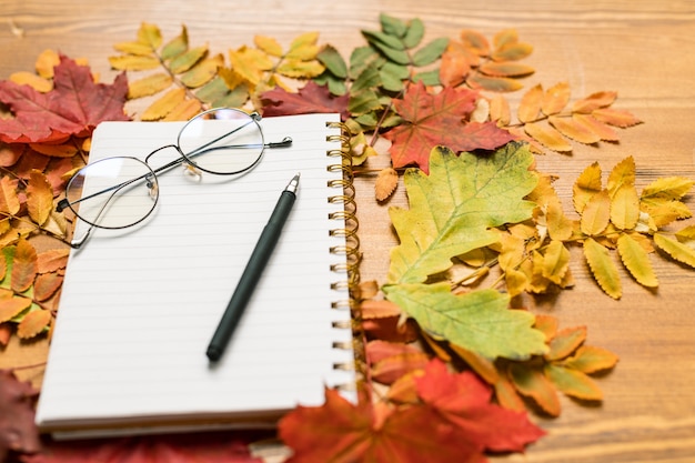 Herfst samenstelling bestaande uit kleurrijke bladeren rond voorbeeldenboek met pen en bril op houten tafel