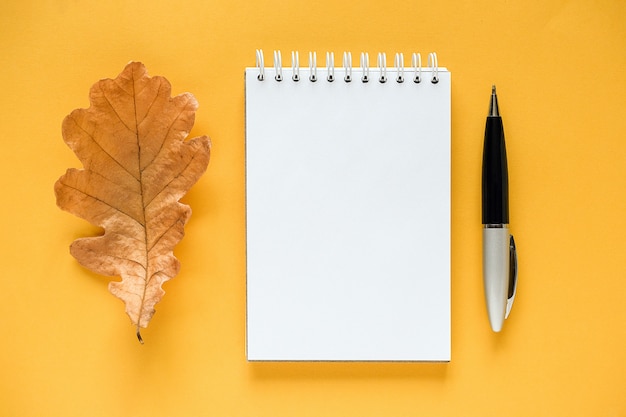 Foto herfst samenstelling. witte lege kladblok, gedroogd oranje eikenblad en pen op geel