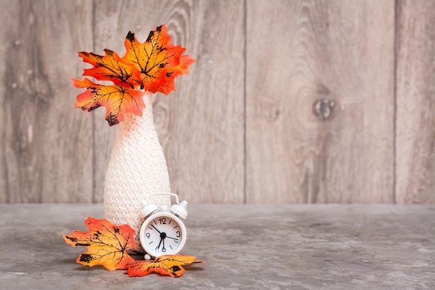 Herfst stilleven. Een vaas met oranje esdoornbladeren en een witte wekker staan op de tafel. Wit-oranje-beige kleurenschema. Kopieer ruimte