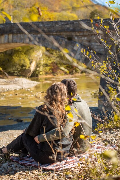 Herfst wandeling. Achteraanzicht. van paar zit op de oever van de rivier. Man en vrouw in warme kleding kijken naar een herfstuitzicht