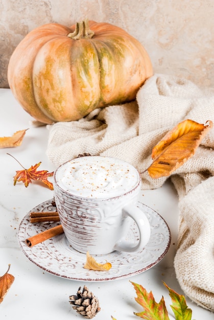 Herfst warme dranken. Pompoen latte met slagroom, kaneelanijs op een witte marmeren tafel, met een trui, herfstbladeren en dennenappels.