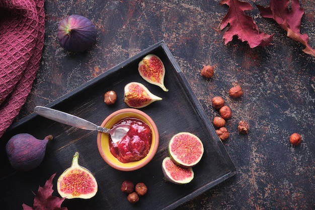 Herfstachtergrond met vijgenjam, lepelnoten en vers gehalveerd vijgenfruit op zwart houten dienblad Donkerbruine achtergrond met droge herfstbladeren