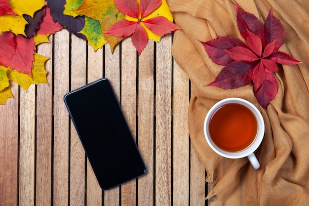 Herfstbladeren, kopje thee, lege zwarte smartphonescherm op tafel, houten achtergrond