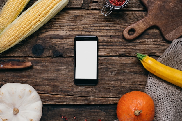 Herfstgroenten: mobiele telefoon met wit leeg scherm, pompoenen en maïs met gele bladeren op hout