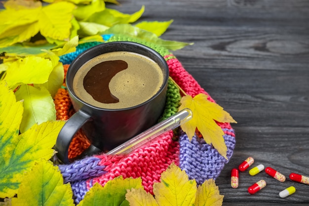 Herfststilleven: een kop warme koffie en een thermometer gewikkeld in een warme sjaal, gele bladeren.