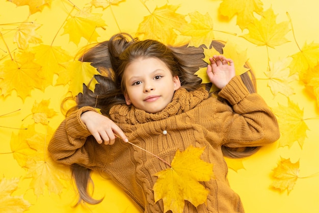 Herfsttijd Een klein meisje in een gebreide trui ligt op een gele achtergrond met gevallen esdoornbladeren