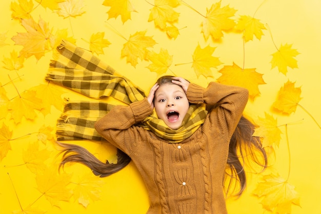 Herfsttijd Een klein meisje ligt op een gele achtergrond tussen gevallen esdoornbladeren in de herfst Het kind, gekleed in een warme gebreide trui en sjaal, greep zijn hoofd vast