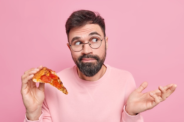 Hesitant bearded young man shrugs shoulders holds tasty pizza being unaware looks clueless away dressed in casual clothes 