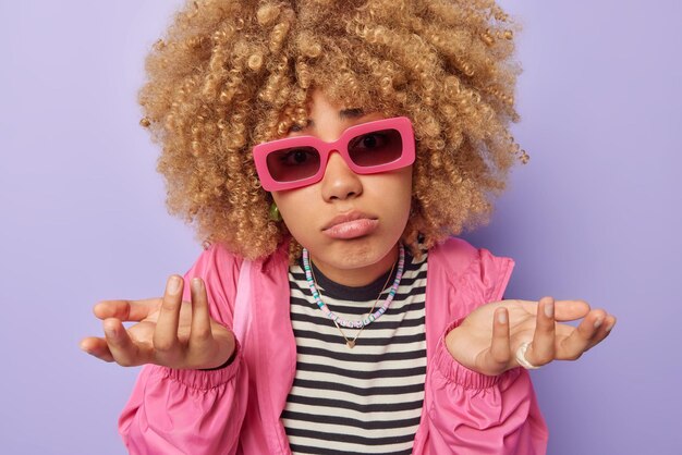 Hesitant clueless woman with curly bushy hair spreads palms\
shrugs shoulders feels unaware doesnt know what to do wears\
sunglasses striped jumper and windbreaker isolated over purple\
background