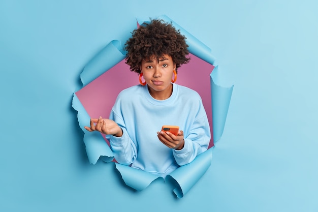 Hesitant unaware Afro American woman doesnt know why her phone isnt working cannot solve problem with modern gadget looks clueless at front dressed casually poses in ripped blue paper wall