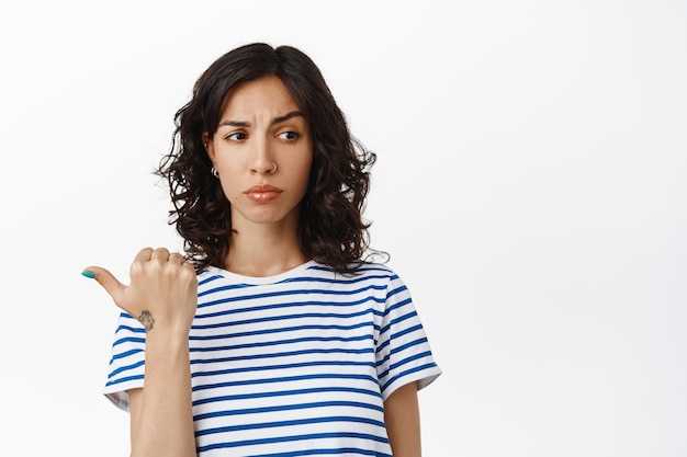 Hesitant and unsure brunette woman, pointing finger left and looking aside, cant decide, not sure about product, feeling suspicious about advertisement, standing over white background