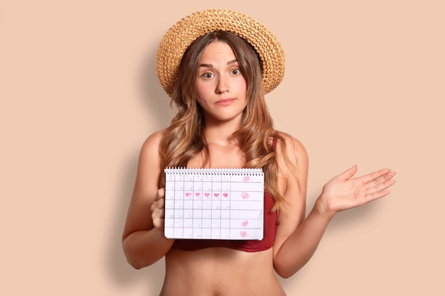 Hesitant young female has vacation abroad, holds period calendar, wonders why she has not regular menstruation, wears stylish straw hat, isolated over studio wall. Women health concept.