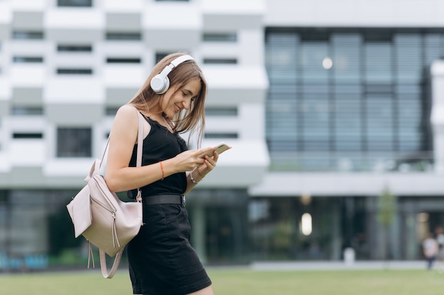 Het aantrekkelijke meisje luistert muziek van telefoon in hoofdtelefoons, typt berichten, terwijl het lopen op de stedelijke straat. Lifestyle.