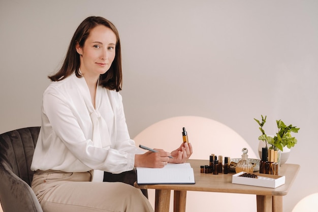 Het aromatherapeut-meisje zit in haar kantoor en houdt een fles aromatische olie in haar handen en schrijft iets op, er liggen essentiële oliën op tafel