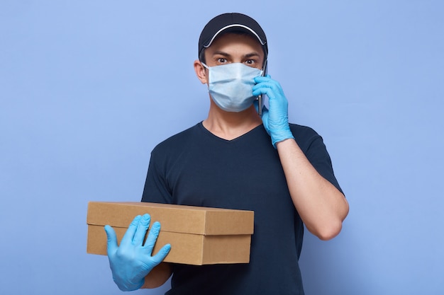 Het beeld van de bezorger die werknemer GLB, t-shirt, masker en handschoenen dragen, houdt bruine kartondoos, kijkt verbaasd, spreekt telefonisch tegen blauwe muur.