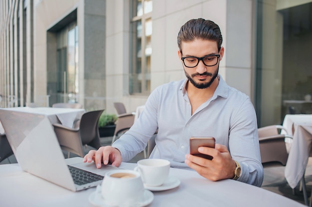 Het beeld van zakenman zit bij lijst en houdt telefoon. Hij kijkt ernaar. Guy heeft twee kopjes koffie en laptop aan tafel. Hij heeft wat rust.