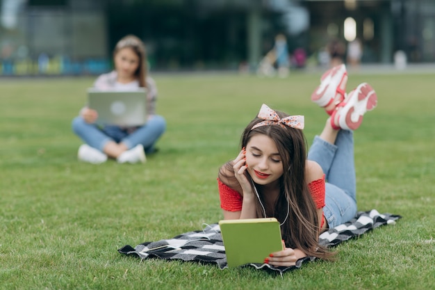 Het boek van de meisjeslezing in park, zittend op gras en het hebben van rust op universitaire campus. Vrouw het luisteren muziek door hoofdtelefoon en het lezen van boek in park