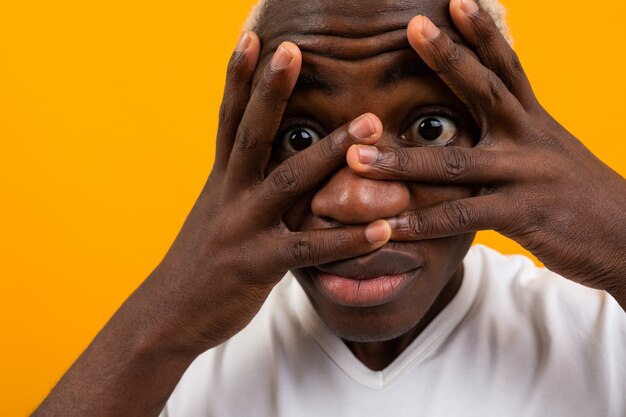 Het close-upportret van een blonde knappe Afrikaanse zwarte mens die zijn gezicht met zijn handen in dient dient een wit T-shirt op geel in