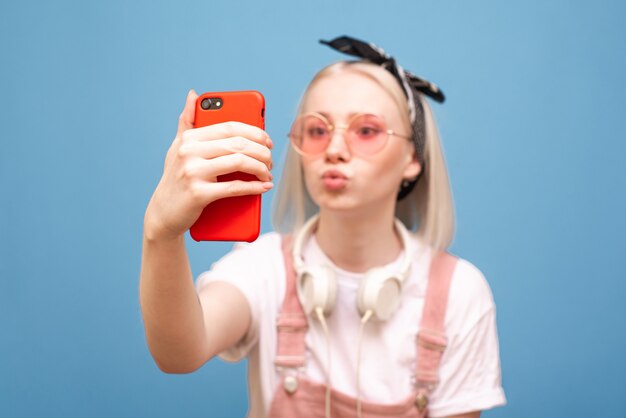 Het close-upportret van een grappig meisje in heldere roze glazen neemt een selfie op een smartphone met een grappig gezicht op een blauwe achtergrond