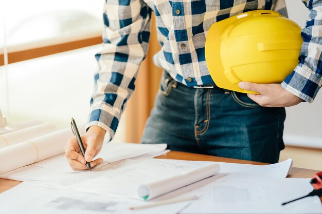 Het concept van de bouw van een architect of ingenieur met een pen die een projectontwerp op een blauwdruk in de hand houdt een gele helm op de werkplek hij droeg een formeel pak