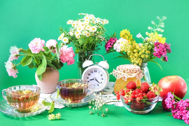 Het concept van een goede zomerochtend kopjes thee onder boeketten van wilde bloemen aardbeien in een kopje potten jam appels wekker groene achtergrond zijaanzicht