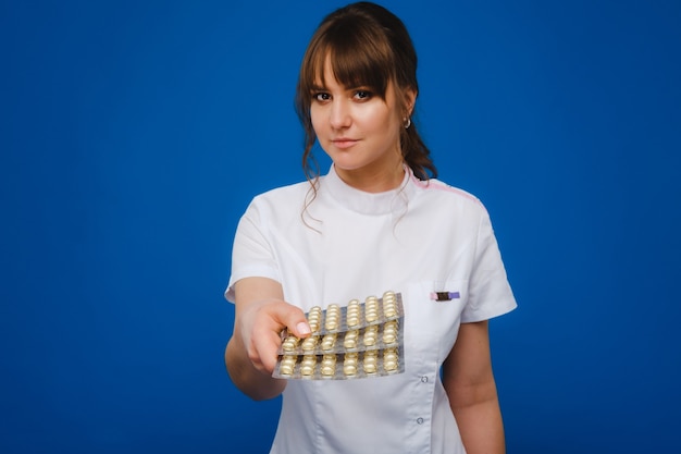 Het concept van gezondheidszorg. Een jonge brunette arts in een witte jas op een blauwe achtergrond toont borden met capsules om in te nemen.